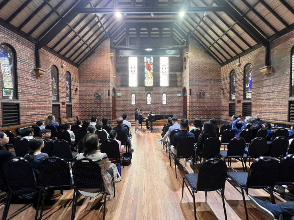 Student violin performance with piano before a full recital audience