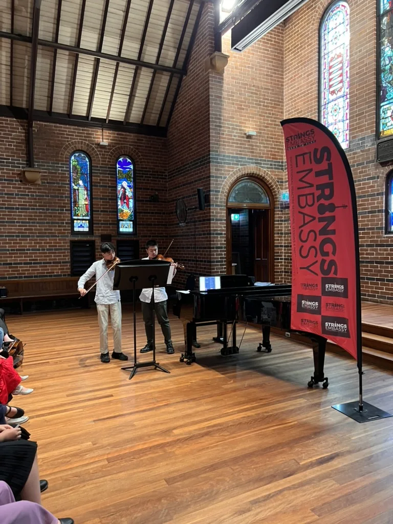 Two young violinists performing together beside a grand piano